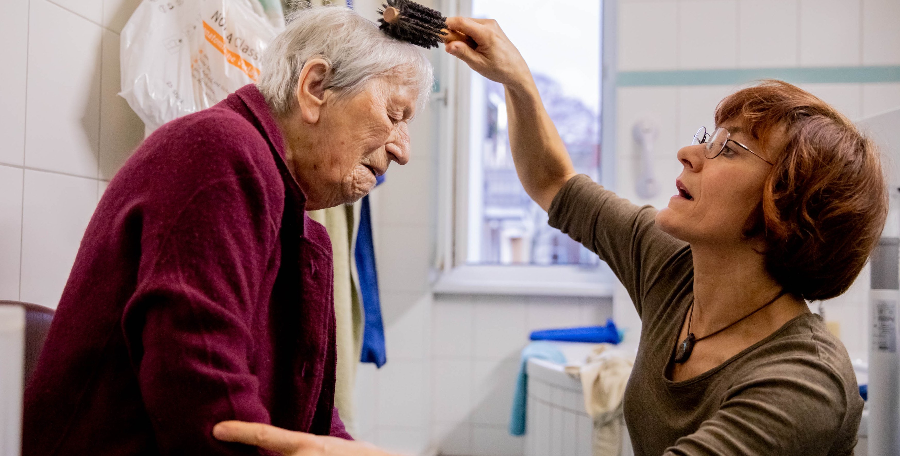 Das Foto zeigt eine &auml;ltere, sitzende Frau, deren Haare von einer mittelalten Frag geb&uuml;rstet werden